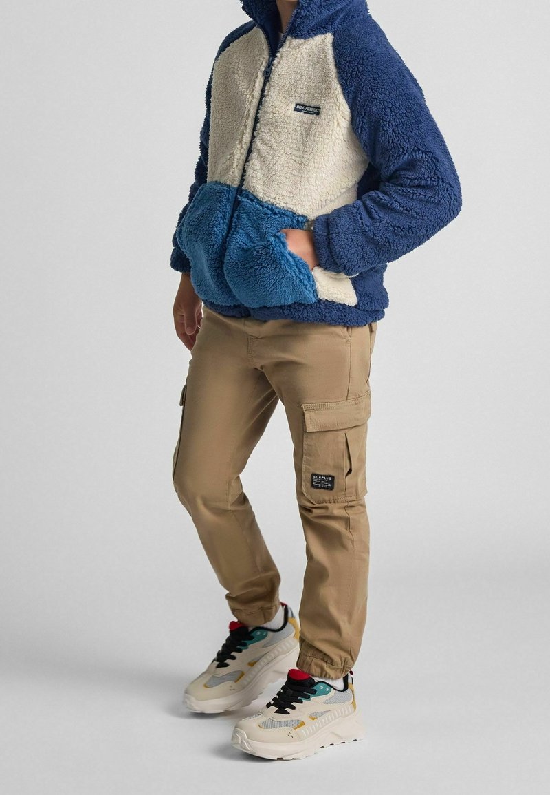 Veste zippée en polaire bleu, crème et bleu clair ; pantalon cargo beige avec poches ; baskets blanches avec accents noirs et colorés.