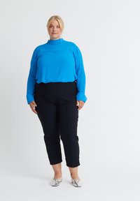 Frau steht mit den Armen seitlich am Körper, trägt ein hellblaues langärmeliges Oberteil, schwarze knöchellange Hose und silberne Ballerinas vor weißem Hintergrund.