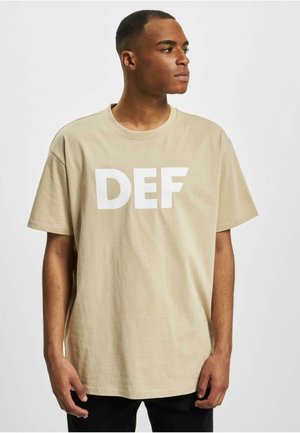 Beige bomull t-skjorte med en hvit "DEF" grafikk på fronten, rund hals design og korte ermer, brukt med svarte bukser.