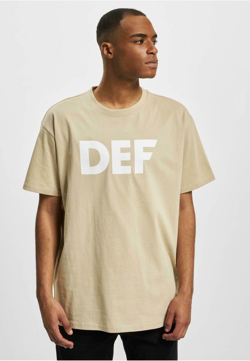 Beige bomull t-skjorte med en hvit "DEF" grafikk på fronten, rund hals design og korte ermer, brukt med svarte bukser.