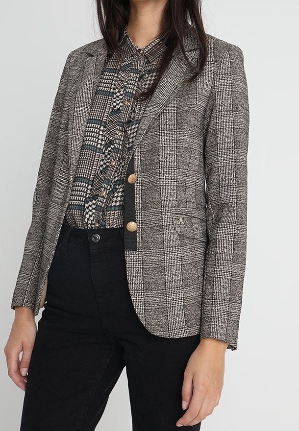 Femme portant un blazer à carreaux gris avec des boutons dorés par-dessus un chemisier noir et blanc à motifs et un pantalon noir, debout devant un fond uni.