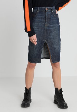 Femme portant une jupe en denim foncé jusqu'aux genoux avec une fente frontale, des bottes de combat noires et un pull noir avec des rayures orange sur les manches.