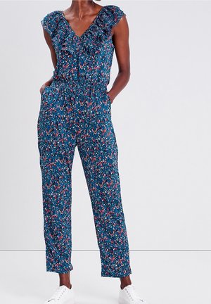 Femme portant une combinaison sans manches bleue à motif floral avec un col à volants et des baskets blanches, debout les mains dans les poches.