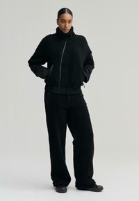 Fekete polárdzseki magas gallérral és cipzárral, mely hozzá illő polárnadrággal van párosítva. Mindkettő puha tapintású és laza fazonú, zsebekkel rendelkezik.