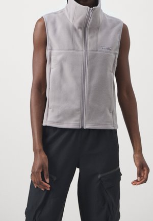 Ärmellose, hellgraue Fleece-Weste mit Reißverschluss und hohem Kragen, getragen über schwarzen Cargohosen, mit Under Armour-Logo auf der Brust.