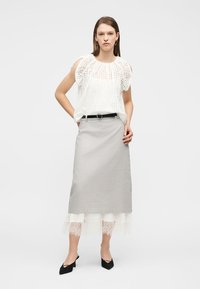 Femme debout les mains dans les poches, portant un chemisier sans manches en broderie anglaise blanche, une jupe midi grise avec un ourlet en dentelle, une ceinture noire et des talons noirs.