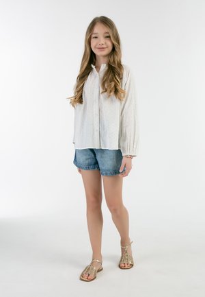 Witte lange mouwen blouse met stippen, gecombineerd met blauwe denim shorts en gouden sandaaltjes met bandjes. Eenvoudige, casual outfit.