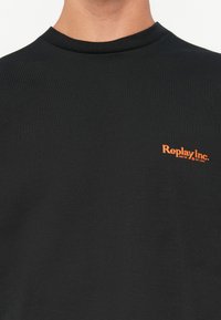Black crew neck t-shirt with small orange text "Replay Inc. REG.TM EST.1981" on the left chest worn by a person.
