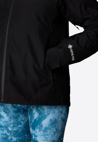 Czarna kurtka Gore-Tex z zamkiem z przodu, regulowanymi mankietami oraz teksturowanym wykończeniem, w połączeniu z legginsami w niebiesko-białe marmurkowe wzory.