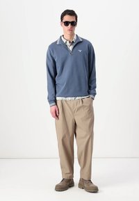 Ανοιχτό μπλε φούτερ από φλις με άσπρη ριγέ κολάρο, που έχει φορεθεί πάνω από πουκάμισο με καρό σχέδιο. Φοράει μπεζ φαρδιά παντελόνια και καφέ μποτάκια μέχρι τον αστράγαλο.