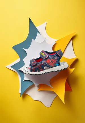Sandalia infantil azul y roja con diseño de Spider-Man, correas de Velcro y suela de goma, presentada sobre un vibrante fondo amarillo de papel.