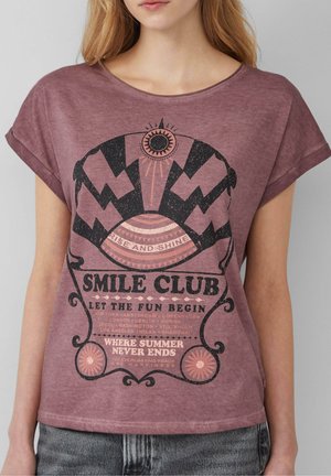 Vrouw draagt een paarse t-shirt met zwarte en roze grafische tekst: "Smile Club", "Rise and Shine" en "Where Summer Never Ends."