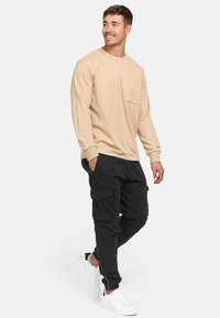 Beige langærmet skjorte med lomme og logo på kraven, parret med sorte cargo bukser med flere lommer og tætsiddende manchetter. Hvide sneakers.