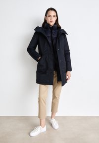 Mørk marineblå dunjakke med avtagbar hette, glidelås og sidelommer. Kombinert med beige cropped bukser og hvite joggesko.