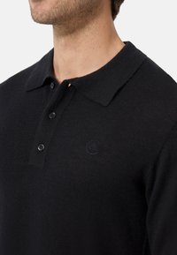 Svart polo skjorta gjord av stickad tyg, med krage, knappslå och en subtil logotyp på bröstet. Slät textur och klassisk design.
