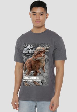 Mann trägt graues Jurassic World T-Shirt mit einem Stygimoloch-Dinosaurier mit Namen und Eigenschaften auf hellem Hintergrund.