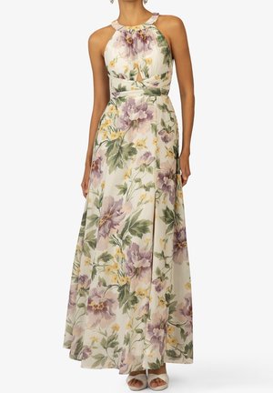 Woman wearing a sleeveless, floor-length floral dress with purple, yellow flowers and green leaves, paired with white open-toe heels.