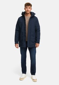 Marine ytterjakke med hette, med glidelås og trykknapper, brukt over en lys genser, kombinert med mørke jeans og hvite joggesko.