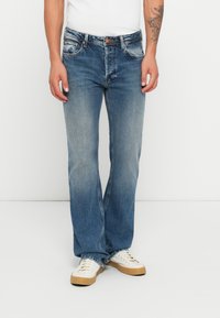 Mann som har på seg falmete blå straight-leg jeans, hvite sneakers med brune såler, og en hvit skjorte, står foran en hvit bakgrunn.