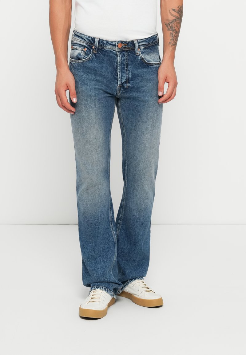 Mann som har på seg falmete blå straight-leg jeans, hvite sneakers med brune såler, og en hvit skjorte, står foran en hvit bakgrunn.