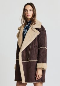 Manteau en suédine marron avec un col et des poignets en sherpa beige, doté d'une fermeture éclair diagonale et de lignes de panneaux contrastantes.