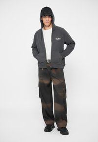 Γκρίζα ζακέτα με φερμουάρ και μπροστινή τσέπη, φορεμένη πάνω από ένα λευκό μπλουζάκι. Παντελόνια cargo με καφέ tie-dye σχέδιο και μαύρα αθλητικά παπούτσια. Μαύρο καπέλο.