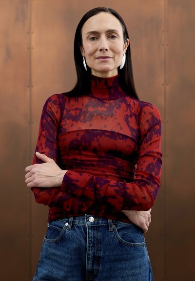 Haut à col roulé transparent à motifs rouges et violets, associé à un jean taille haute en denim. Des boucles d'oreilles en argent simples complètent la tenue.