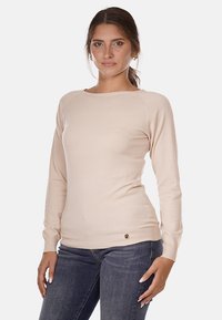 Maglione beige a maniche lunghe con scollo rotondo, texture liscia, vestibilità slim e sottile logo sul bordo inferiore. Abbinato a jeans scuri.