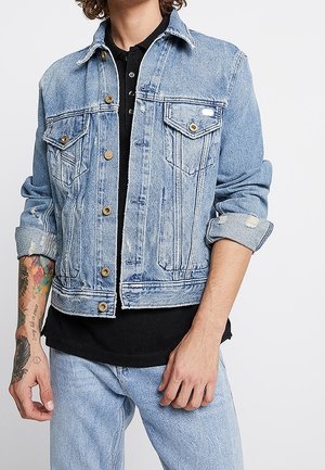 Homme portant une veste en jean bleu clair avec les manches retroussées par-dessus une chemise noire et un jean bleu clair assorti, montrant des tatouages colorés sur les bras.