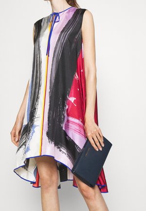 Femme portant une robe sans manches à imprimé abstrait multicolore, tenant une pochette bleu foncé avec des lettres dorées.