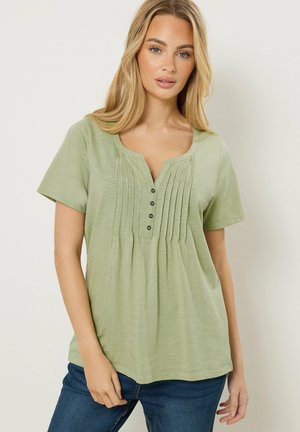 Kobieta w jasnookiej zielonej bluzce z krótkim rękawem i plisowaną, zapinaną na guziki częścią z przodu, w niebieskich dżinsach, stojąca na jednolitym jasnym tle.