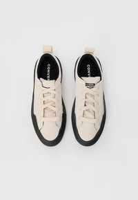 Par tenisice bež boje u niskom modelu marke Converse s crnim gumenenim potplatima i bijelim vezicama, prikazane odozgo na bijeloj pozadini.