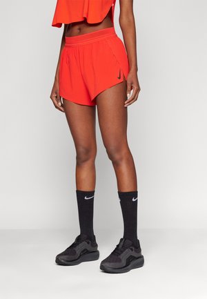 Atletyczna kobieta ubrana w jasnopomarańczowe krótkie spodenki Nike, czarne skarpetki Nike i czarne buty do biegania, stojąca na tle jednolitego jasnego tła.