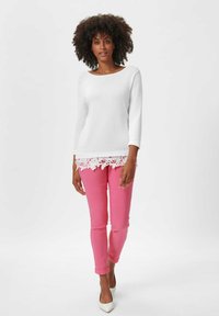 Witte lange mouwen top met bloemenkant aan de onderkant, gecombineerd met aansluitende roze broek en witte puntige hakken. Soepele textuur en eenvoudig ontwerp.
