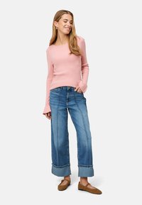 Helles rosa geripptes Langarmshirt, weite blaue Jeanshose mit hochgekrempelten Säumen und braune flache Schuhe, getragen von einer Frau vor einem neutralen Hintergrund.