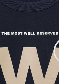 T-shirt in cotone blu navy con testo bianco che recita "IL PIÙ MATO CHE MERITA" sopra un design beige a forma di 'W' e un accento circolare.