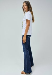 T-shirt en coton blanc avec un imprimé graphique, associé à un jean évasé bleu foncé. Le mannequin se tient de profil, portant des sandales beige.