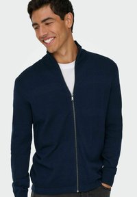 Pull en maille bleu marine à fermeture éclair avec des motifs texturés, col et poignets côtelés, et une fermeture éclair lisse. Porté par-dessus une chemise blanche.