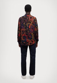Camicia a maniche lunghe con un intricato motivo paisley multicolore su uno sfondo scuro, abbinata a pantaloni in denim scuro e scarpe bianche.