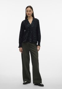 Vrouw staand, gekleed in een zwarte blouse met patroon en lange mouwen, gecombineerd met olijfgroene wijde broekspijpen en zwarte schoenen, tegen een witte achtergrond.