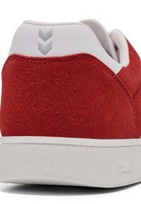 Red suede sneaker featuring a white leather heel and textured rubber sole. Details include three gray chevron accents on the heel.