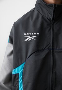 Black and grey jacket featuring teal accents, a high collar, and the "BOTTER" logo in white on the front. Smooth, lightweight fabric.