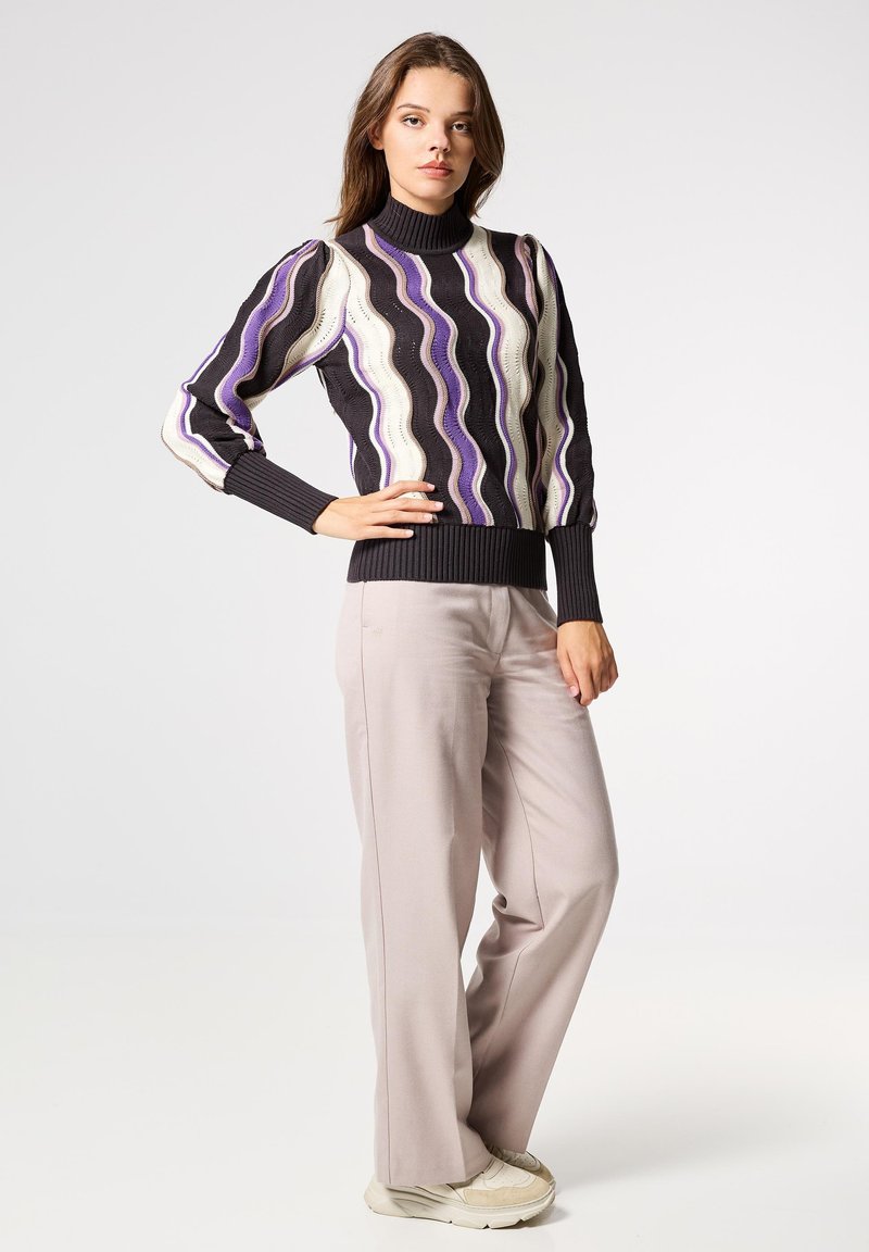 Jersey de punto con patrones ondulados en negro, blanco y púrpura; cuello alto y puños acanalados. Combinado con pantalones gris claro y zapatillas.