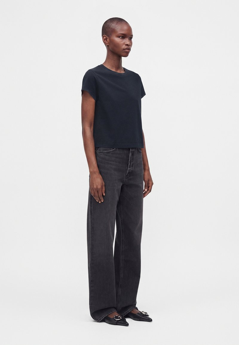 Femme aux cheveux courts portant un t-shirt noir à manches courtes, un jean noir à jambes larges et des ballerines noires pointues avec une boucle argentée.