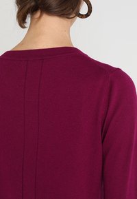 Fuchsia stickad tröja med vid rundad halsringning, ribbad kant och en central sömdetalj på baksidan. Mjuk textur, långa ärmar.