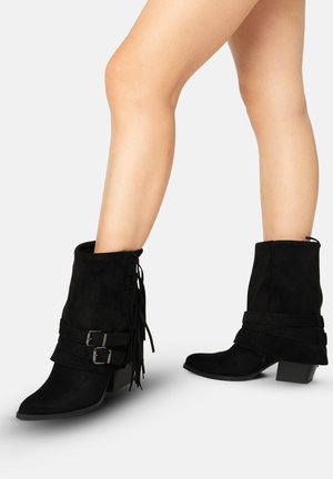 Gambe con stivali di camoscio nero a metà polpaccio con fibbie laterali, dettagli con frange e tacchi bassi a blocchetto su sfondo bianco semplice.