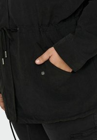 Juodas švarkas su laisvu siluetu, su raišteliu juosmenyje ir šoninėmis kišenėmis. Tekstūruota medžiaga ir matomi siuvimo detalės.