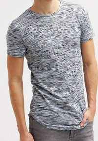 Muž v úzkém, krátkém šedém a černém tečkovaném tričku s krátkým rukávem, s jednou rukou v kapse šedých kalhot.