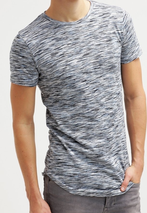 Muž v úzkém, krátkém šedém a černém tečkovaném tričku s krátkým rukávem, s jednou rukou v kapse šedých kalhot.