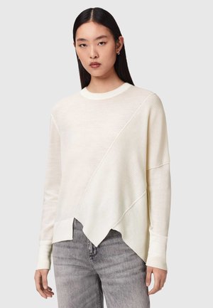 Młoda kobieta z długimi ciemnymi włosami, ubrana w asymetryczny kremowy sweter i jasnoszare dżinsy, stojąca na tle jednolitego jasnego tła.